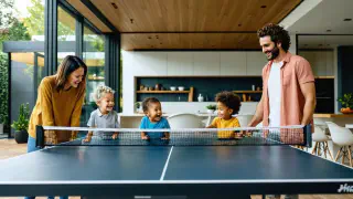 Ein modernes, gut beleuchtetes Zuhause mit einer Familie über drei Generationen, die sich um einen Tischtennistisch versammelt hat, lächelnd und vertieft ins Spiel.